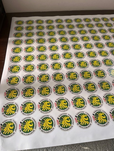 Arkusze z okrągłymi naklejkami z logo 'Gryf Biskupeckie Stowarzyszenie Detektorystów' w kolorach zielonym, żółtym i czarnym, ułożone równo na białym podłożu.