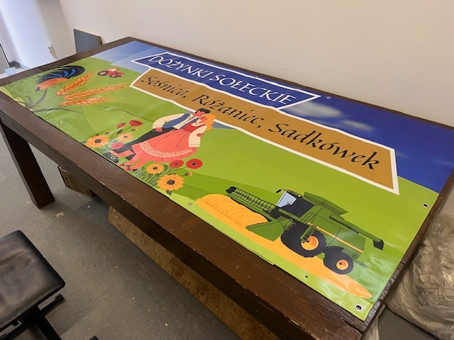 Baner reklamowy na drewnianym stole, z grafiką dożynek Soleckich: kogut, traktor, para taneczna, kombajn. Żywe kolory, widoczne oczka mocujące.