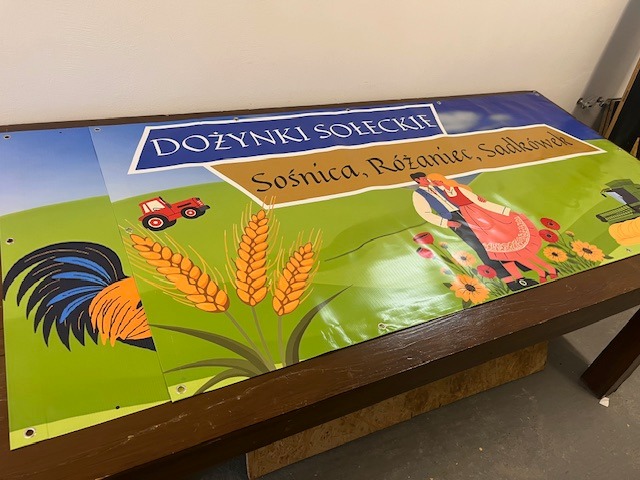 Banery z motywem dożynkowym, z grafikami traktora, zboża i pary w strojach ludowych, leżące na stole. Widoczne otwory do mocowania.