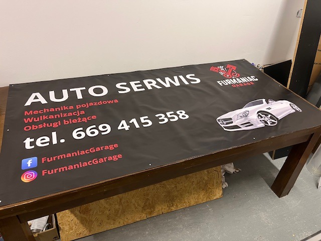 Baner reklamowy warsztatu samochodowego z grafiką auta i danymi kontaktowymi, leżący na stole z ciemnego drewna w pomieszczeniu warsztatowym.