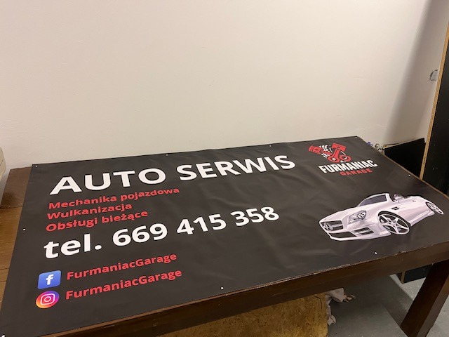 Czarny baner reklamowy auto serwisu z białymi napisami, logotypem i grafiką samochodu, leżący na brązowym stole. Widoczne media społecznościowe i numer telefonu.