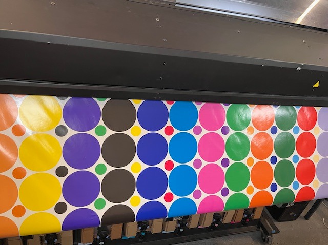 Drukarka wielkoformatowa w trakcie drukowania wzoru z kolorowych kół na białym tle, widoczne kartonowe rolki z materiałem do druku.