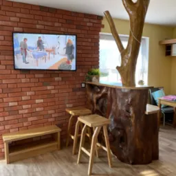 Unikatowy bar z pnia drzewa w salonie z ceglaną ścianą. Dębowe hokery i ławka. Telewizor na ścianie. Nietypowe rozwiązanie aranżacyjne.