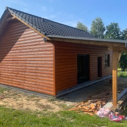 Jakub Spórna - Nowoczesna, brązowa konstrukcja z drewna z czarnym dachem, z elementami otwartej zabudowy, w otoczeniu zieleni i fragmentami materiałów budowlanych.