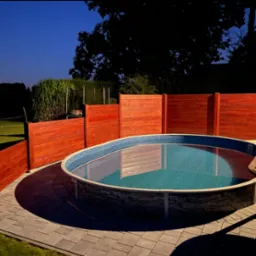 Okazały basen ogrodowy z drewnianym obramowaniem, otoczony kostką brukową i zielenią, sfotografowany w nocy przy stonowanym oświetleniu.