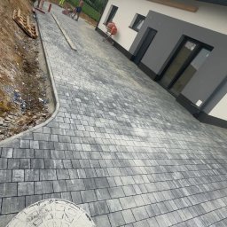 Jakub Spórna - Ułożona szara kostka brukowa prowadząca do nowoczesnego domu z szarymi akcentami. W tle pracownicy i betoniarka. Widoczny właz kanalizacyjny.