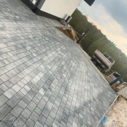 Jakub Spórna - Ułożona szara kostka brukowa na podjeździe domu z szarą elewacją. W tle widoczny samochód ciężarowy i drzewa. Ujęcie z niskiej perspektywy.