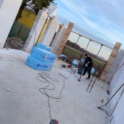 Jakub Spórna - Budowa domu z białych bloczków: pracownik z narzędziem, widoczne materiały izolacyjne i narzędzia. Etap wznoszenia ścian, częściowo zbrojonych.