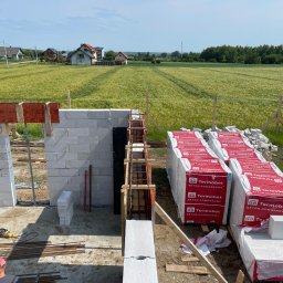Jakub Spórna - Wznoszenie ścian domu z betonu komórkowego Termobet. Widoczne zbrojenia, szalunki i narzędzia. W tle zielone pola i zabudowa wiejska.