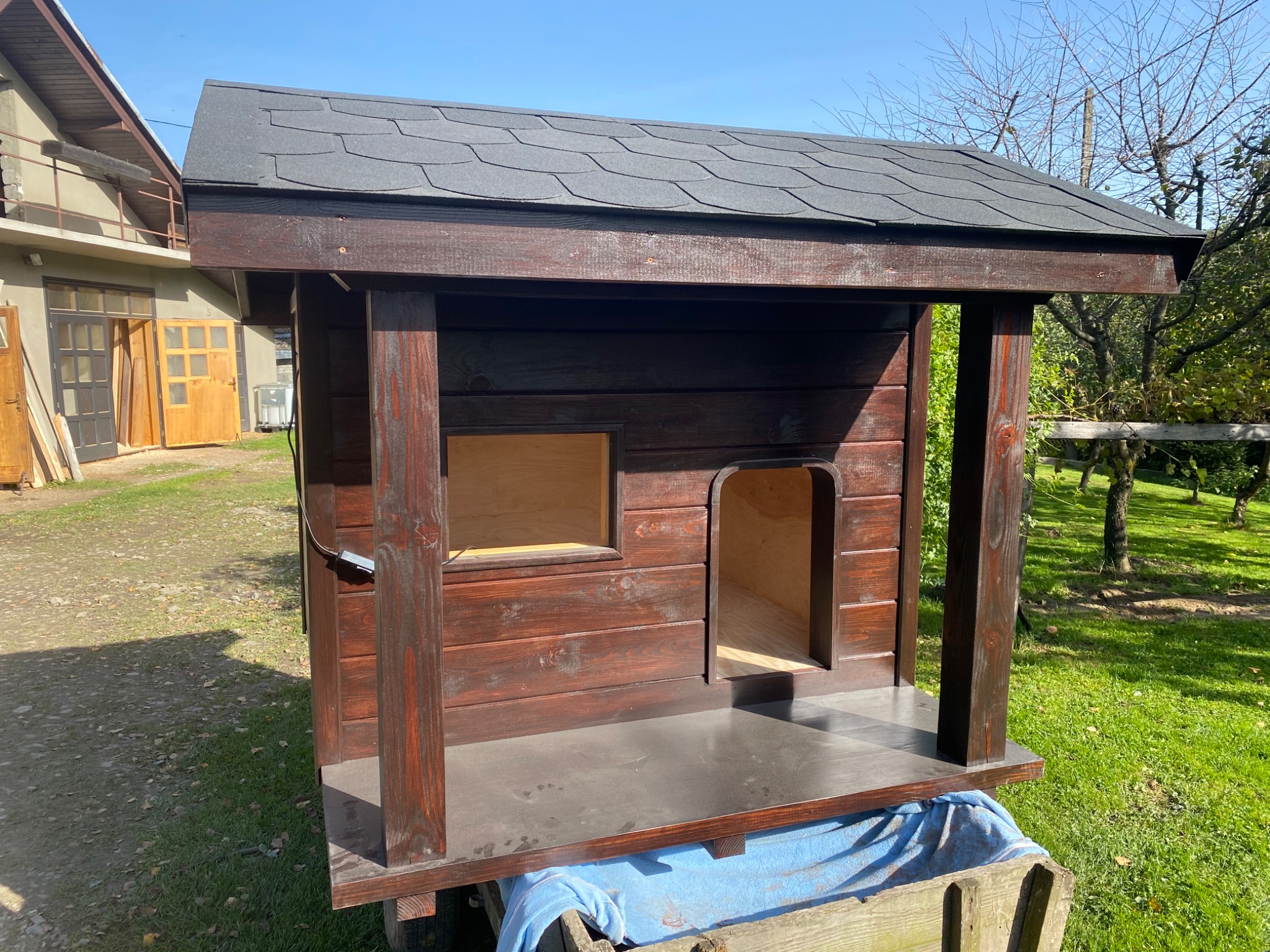 Brązowa, drewniana konstrukcja z dachem z gontu bitumicznego, przypominająca domek, z oknem i wejściem, na tle zieleni i budynku gospodarczego.