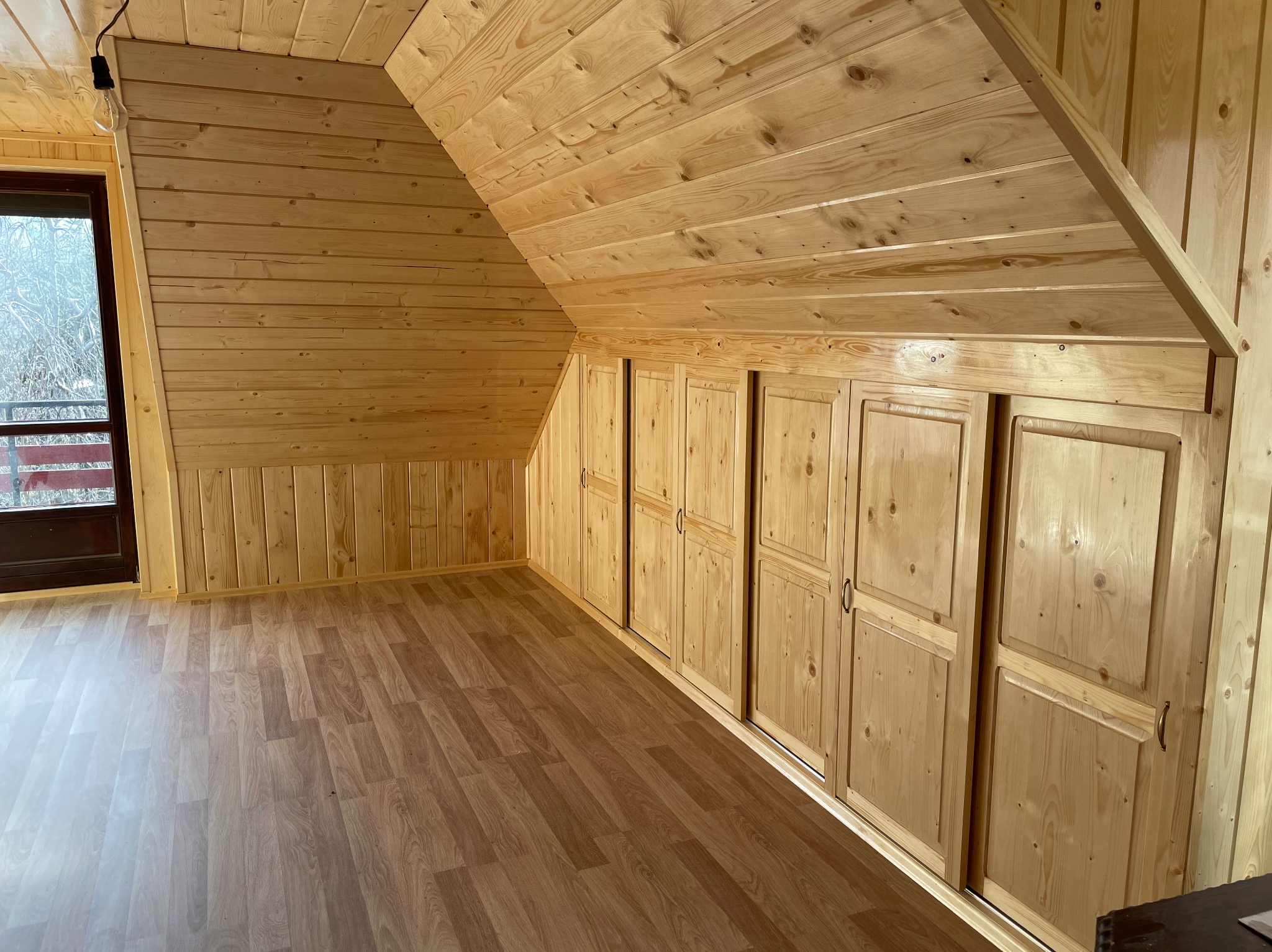 Drewniane wnętrze poddasza z zabudową szaf w skosie, wykończone boazerią. Widoczne parkiety i okno z widokiem na zewnątrz. Surowy klimat drewna.