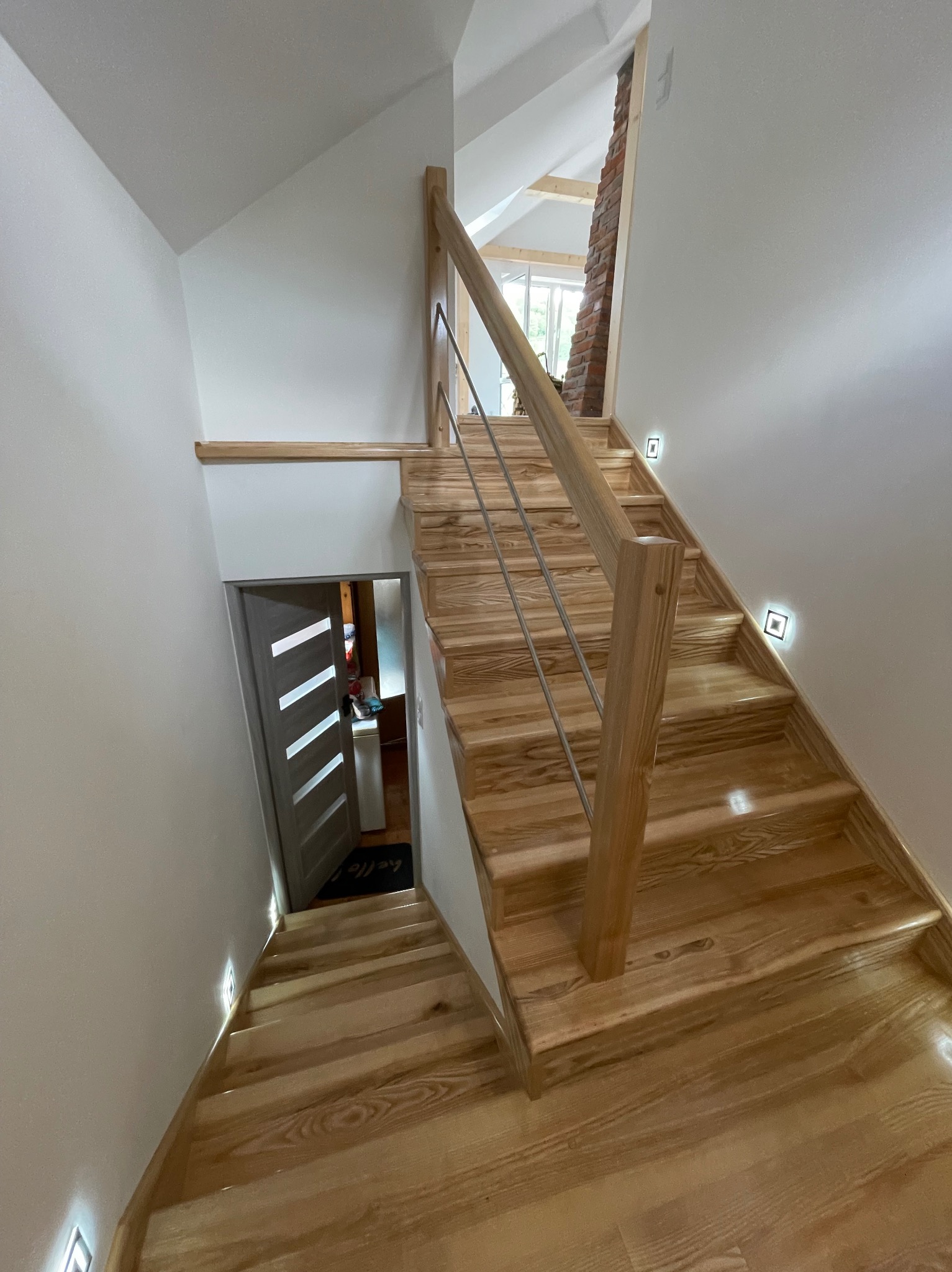 Jasne, drewniane schody z metalową balustradą, oświetlone punktowymi lampkami ściennymi. Widok na otwarte drzwi i fragment wnętrza domu.