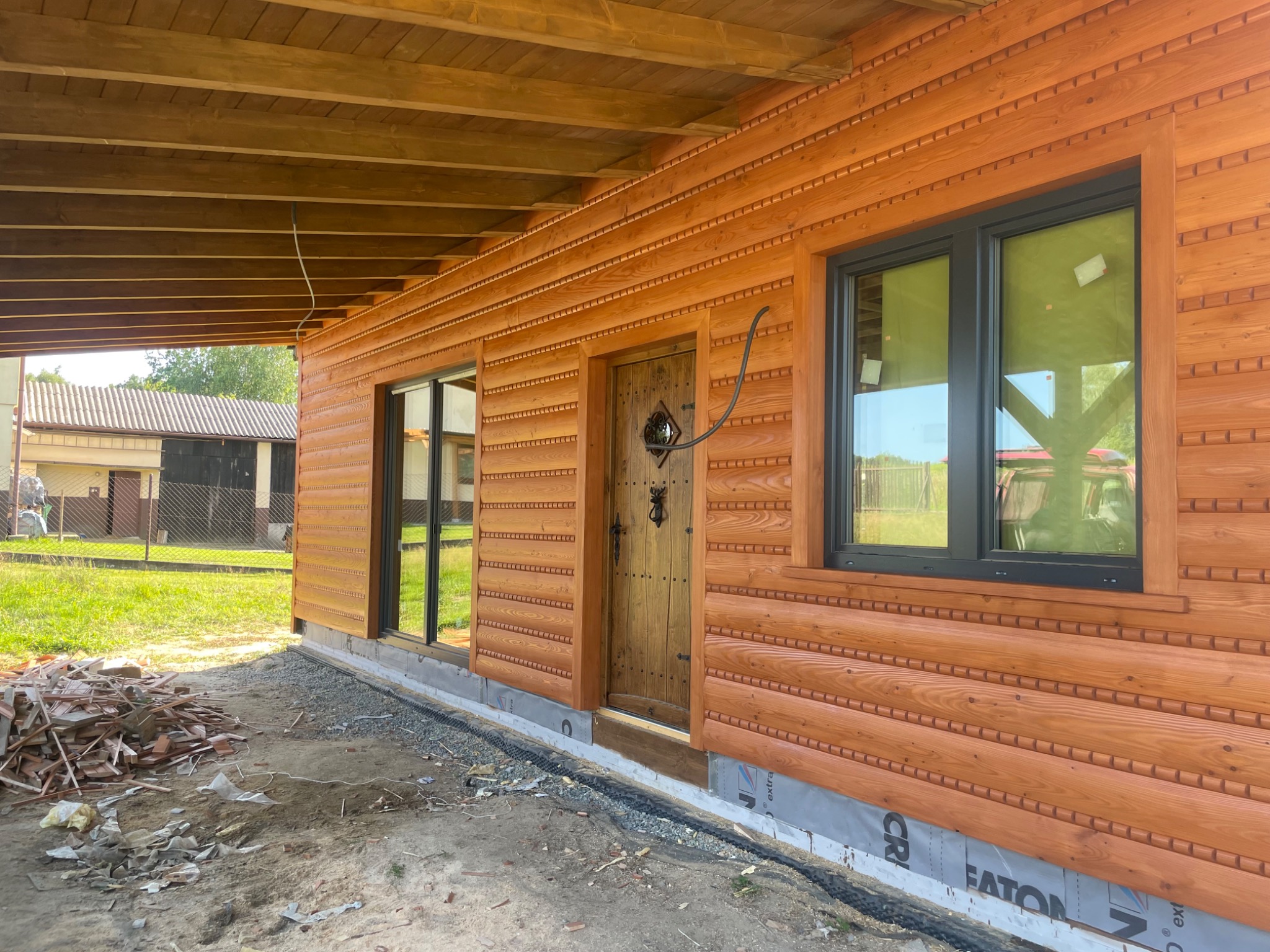 Wykończona drewniana elewacja domu z czarnymi ramami okiennymi i ozdobnymi listwami. Widoczny fragment podbitki dachowej i teren wokół budynku w trakcie prac.