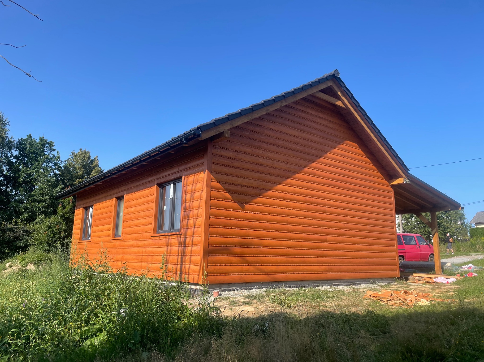 Nowoczesny dom z drewnianą elewacją w odcieniu brązu, z ciemnymi oknami i dachem, na tle błękitnego nieba. Widoczna część zadaszenia i czerwony samochód.