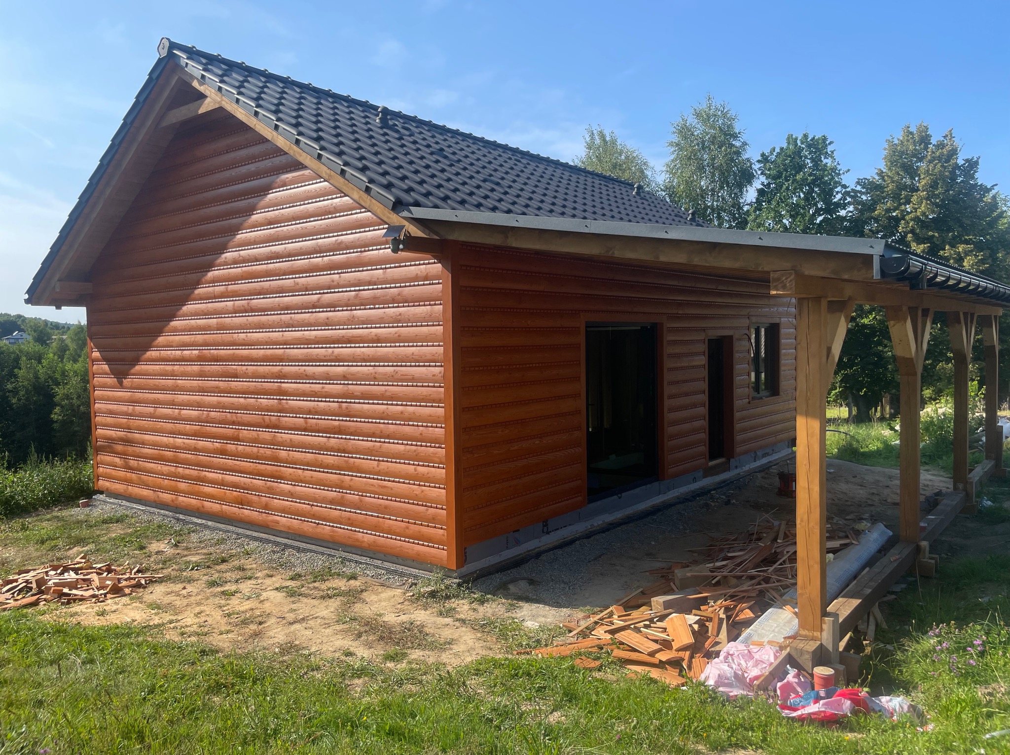 Nowoczesna, brązowa konstrukcja z drewna z czarnym dachem, z elementami otwartej zabudowy, w otoczeniu zieleni i fragmentami materiałów budowlanych.