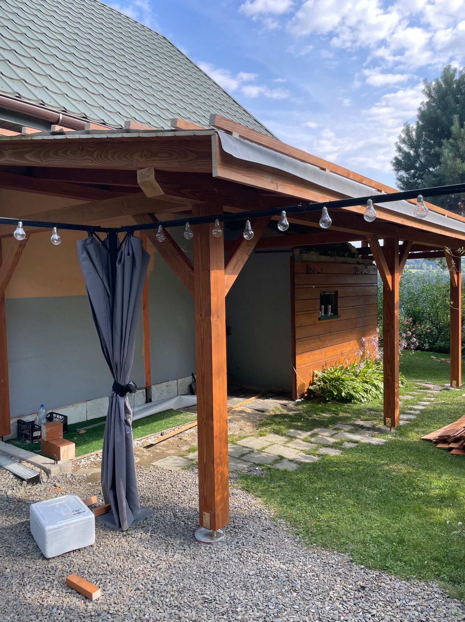 Konstrukcja z drewna z zasłoną i oświetleniem na zewnątrz, z widoczną w tle zielenią i budynkiem. Elementy konstrukcyjne w kolorze ciepłego brązu.