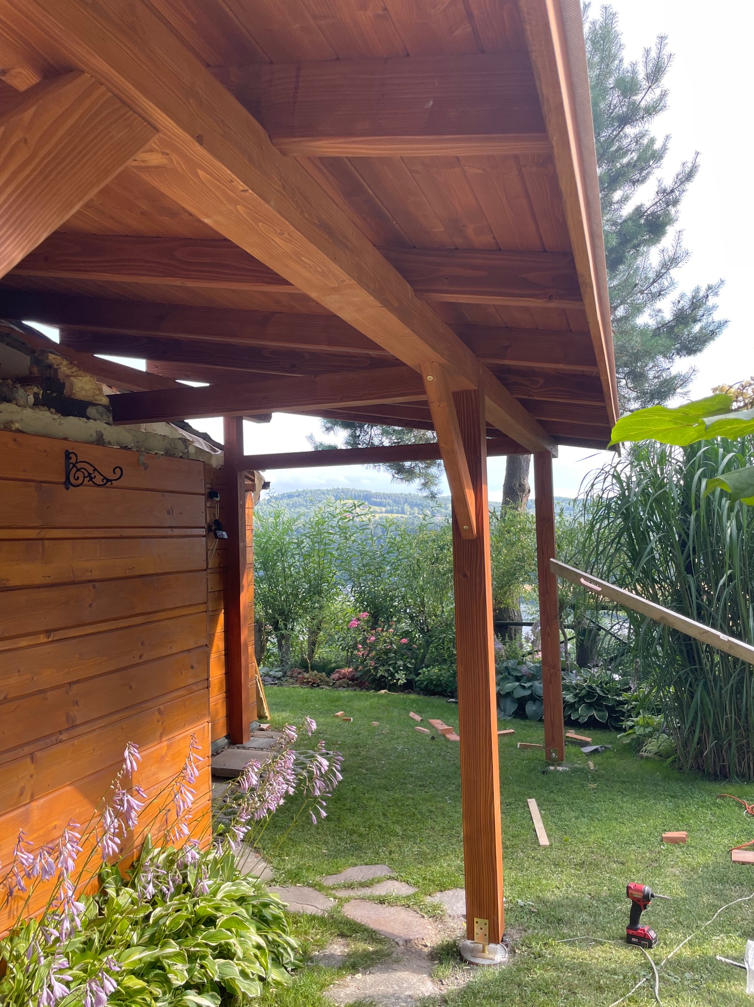Brązowa konstrukcja z drewna na zewnątrz, częściowo zadaszona, z widokiem na zielony ogród i odległe wzgórza w tle. Widać narzędzia i materiały budowlane.