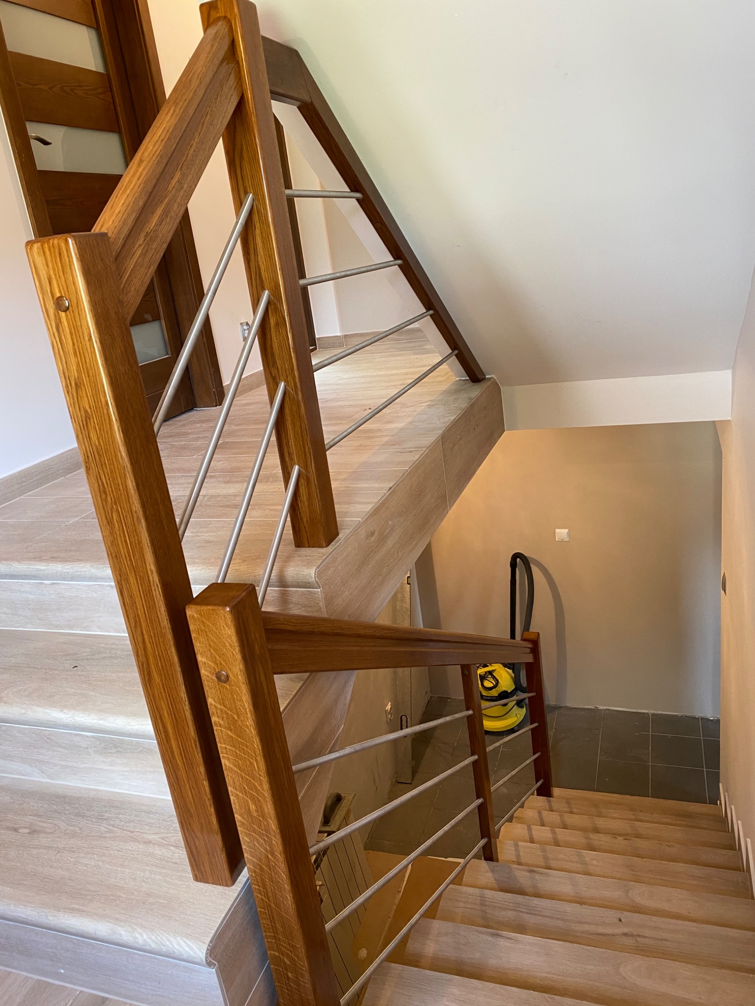 Nowoczesne schody drewniane z metalowymi balustradami, widok z góry. Jasne drewno i neutralne kolory ścian. Na podeście stoi odkurzacz.