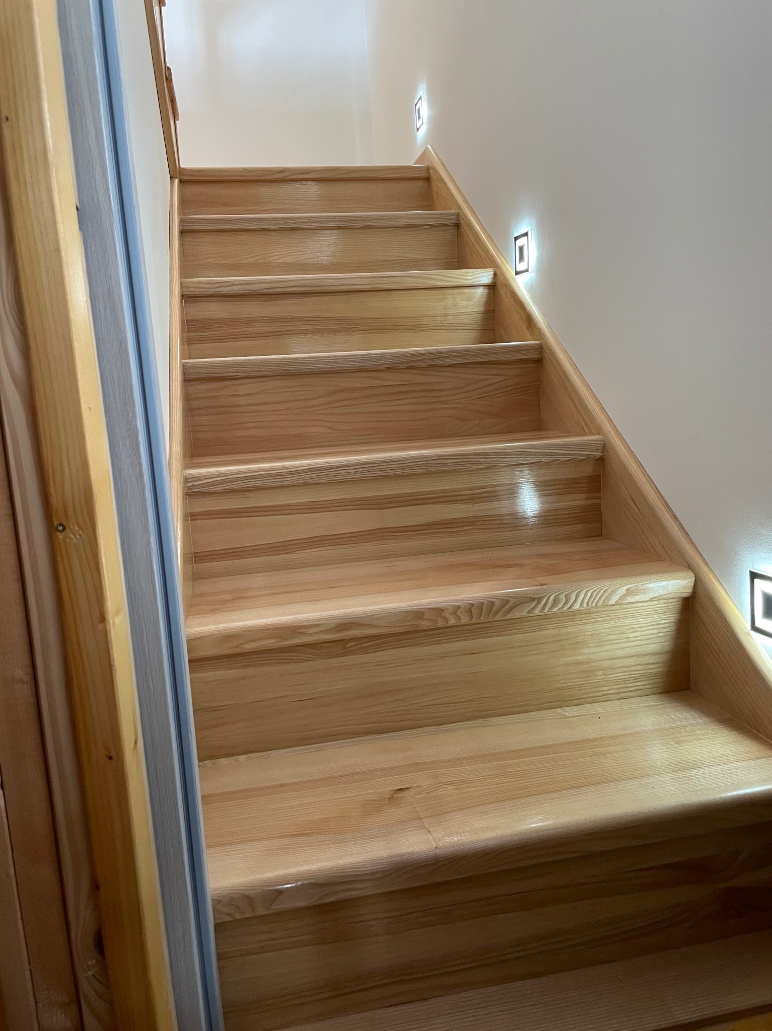 Jasne, drewniane schody z dyskretnym oświetleniem punktowym przy ścianie, widok z dołu, ukazujący solidne wykonanie i naturalny odcień drewna.