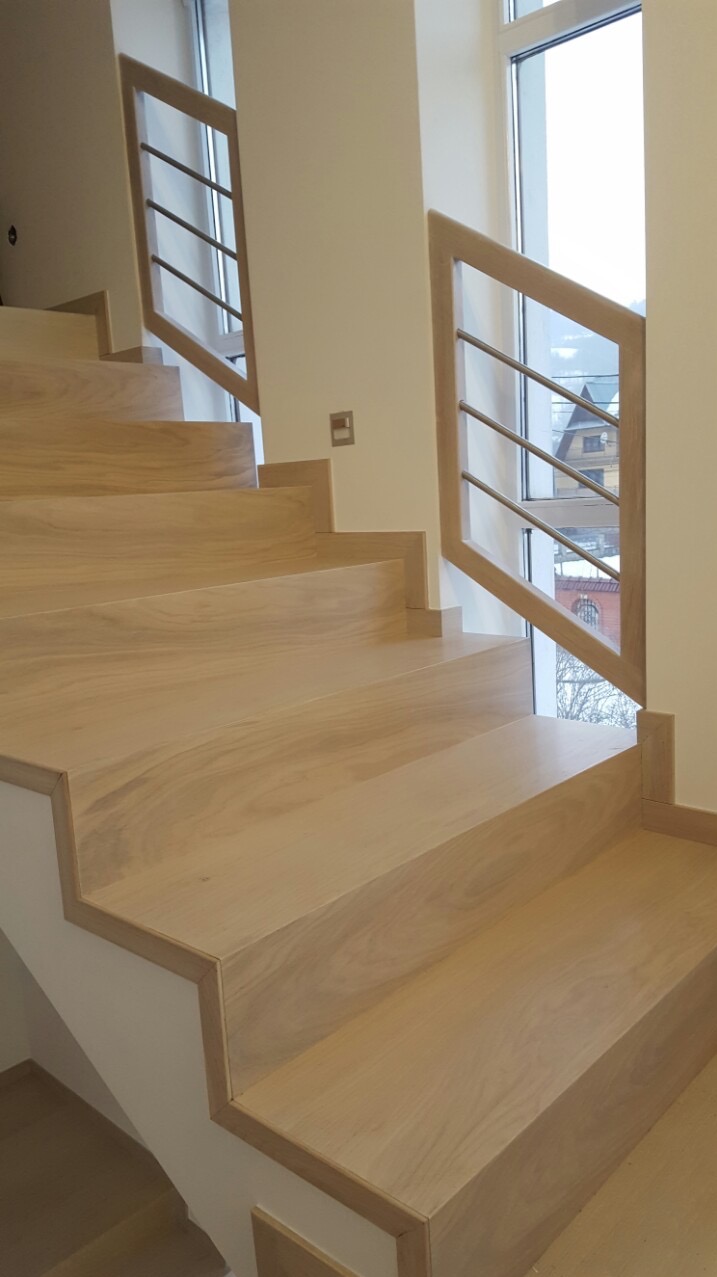 Jasne drewniane schody z nowoczesną balustradą ze stali nierdzewnej, widok z dołu, naturalne oświetlenie z okna w tle. Detal wykończenia.