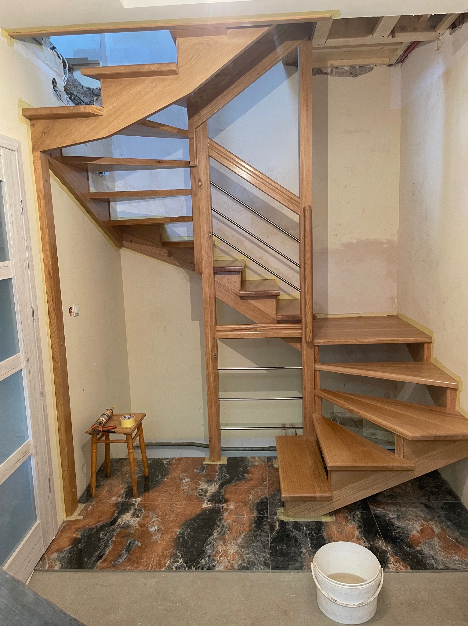 Montaż drewnianych schodów z metalową balustradą w jasnym wnętrzu. Widoczne narzędzia i fragment podłogi. Etap prac wykończeniowych.