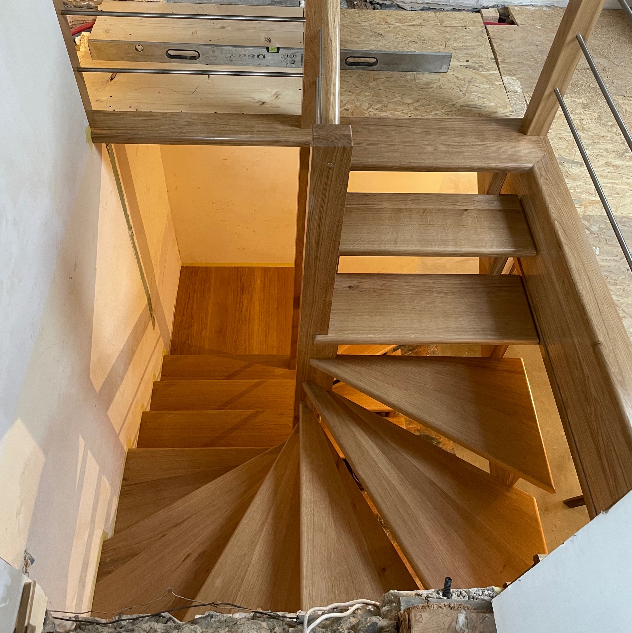 Kręte, dębowe schody z metalową balustradą w trakcie montażu, widok z góry. Na podłodze poziomica. Ściany w trakcie wykończenia.