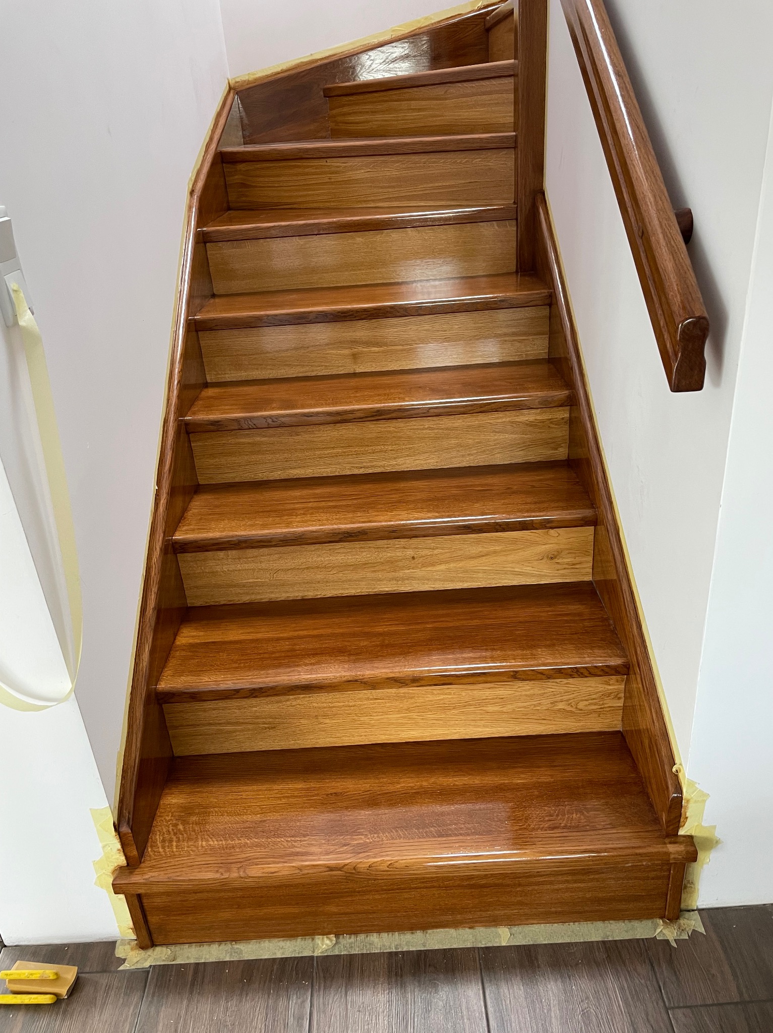 Polakierowane drewniane schody z poręczą, zabezpieczone taśmą malarską przy ścianie. Widoczne słoje drewna i połysk lakieru. Ujęcie od dołu.