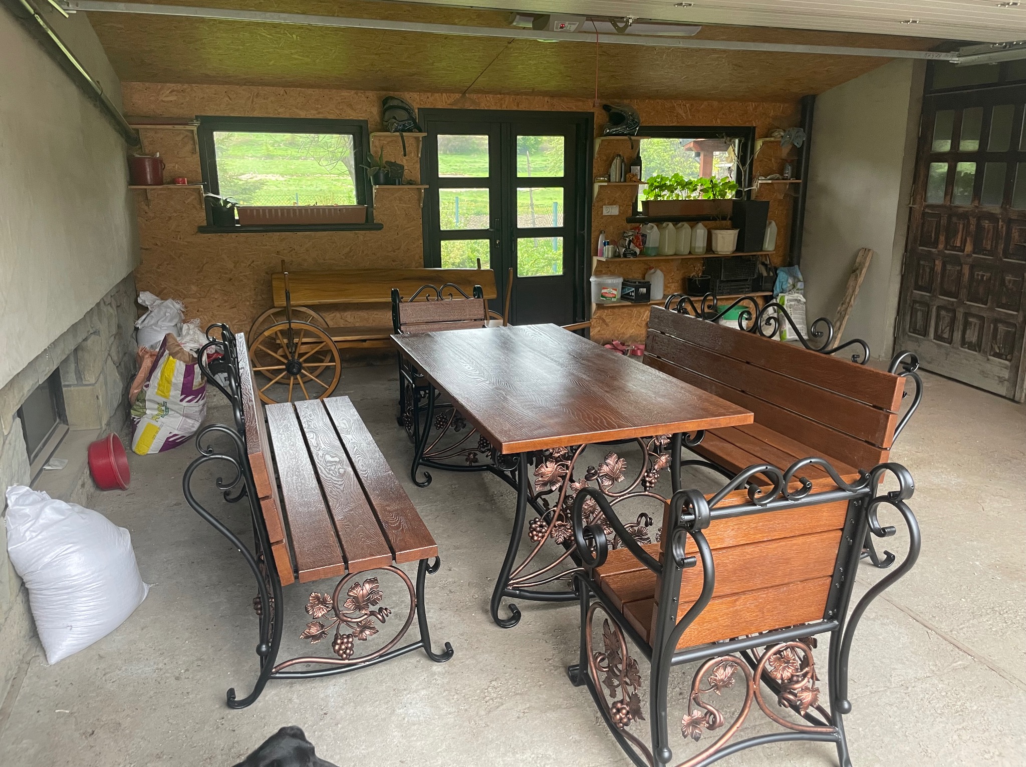 Rustykalny stół i ławki z kutymi elementami w motywie winogron, ustawione w zadaszonym pomieszczeniu z drewnianą boazerią i oknami z widokiem na zieleń.