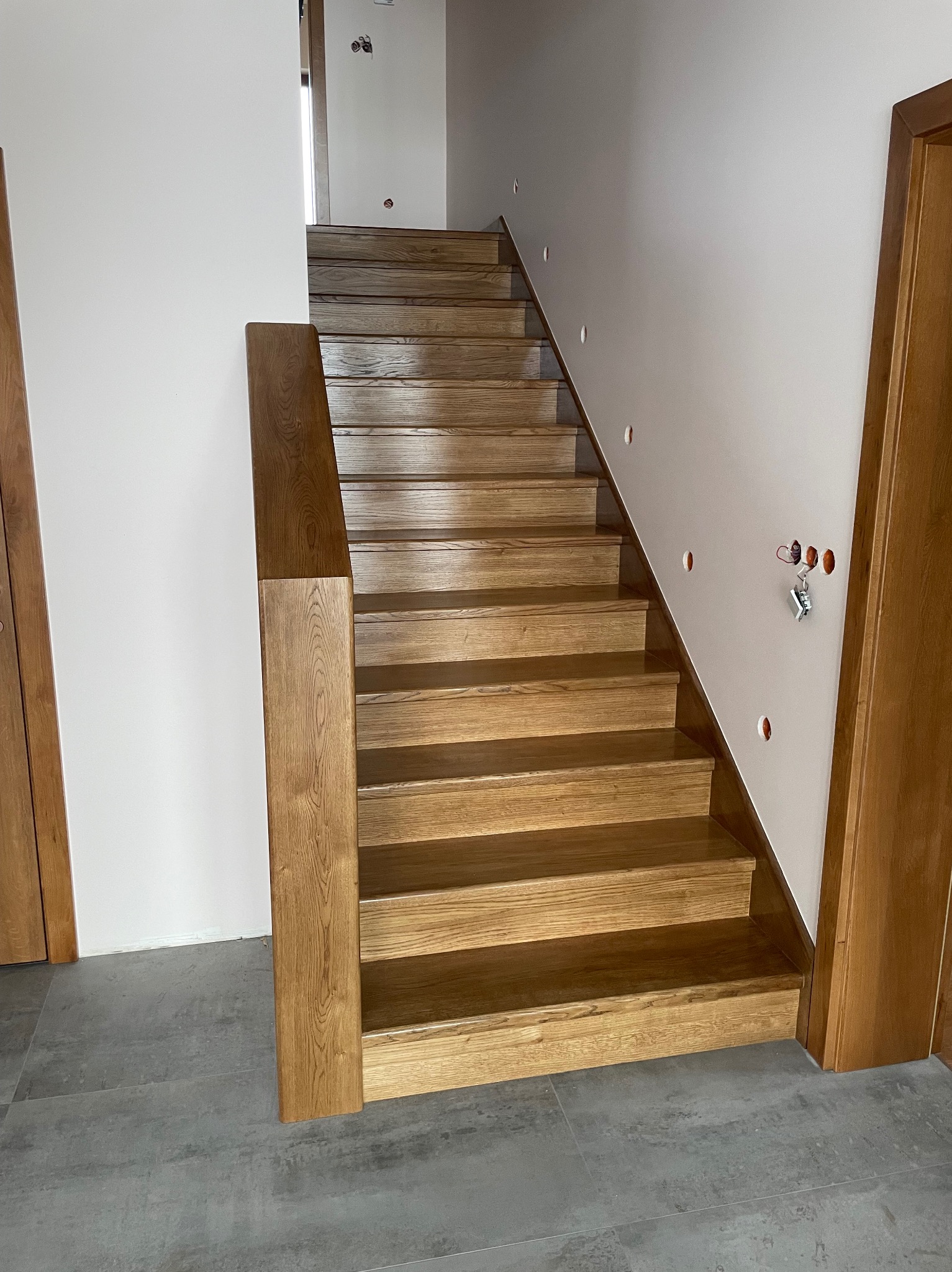 Dębowe schody z podestem, widok z dołu. Naturalne wykończenie drewna, minimalistyczne białe ściany z przygotowanymi punktami oświetleniowymi. Szare płytki podłogowe.