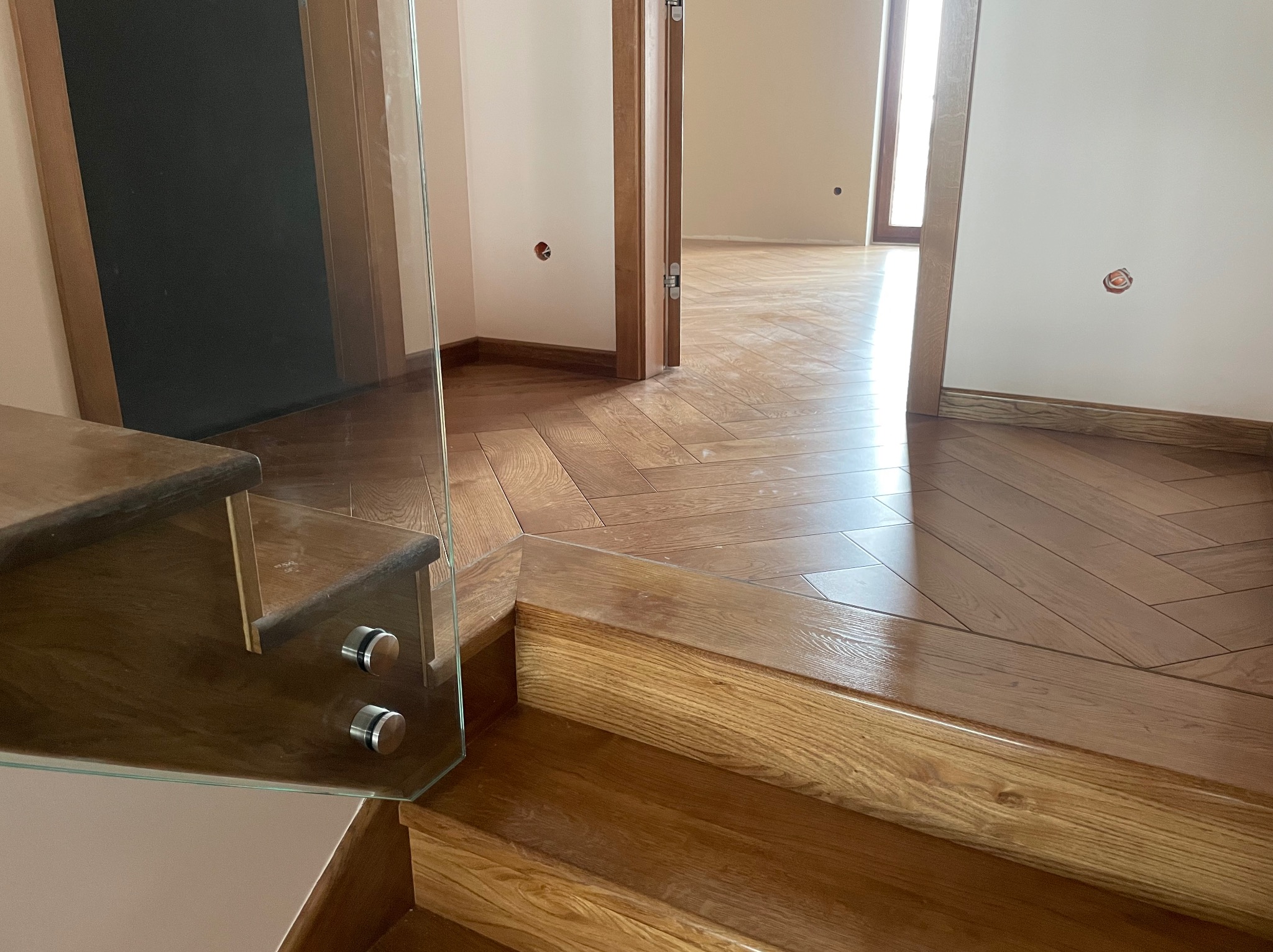 Nowoczesne schody drewniane z balustradą szklaną i parkietem ułożonym w jodełkę. Widok z dołu, jasne wnętrze. Dbałość o detal i wykończenie.