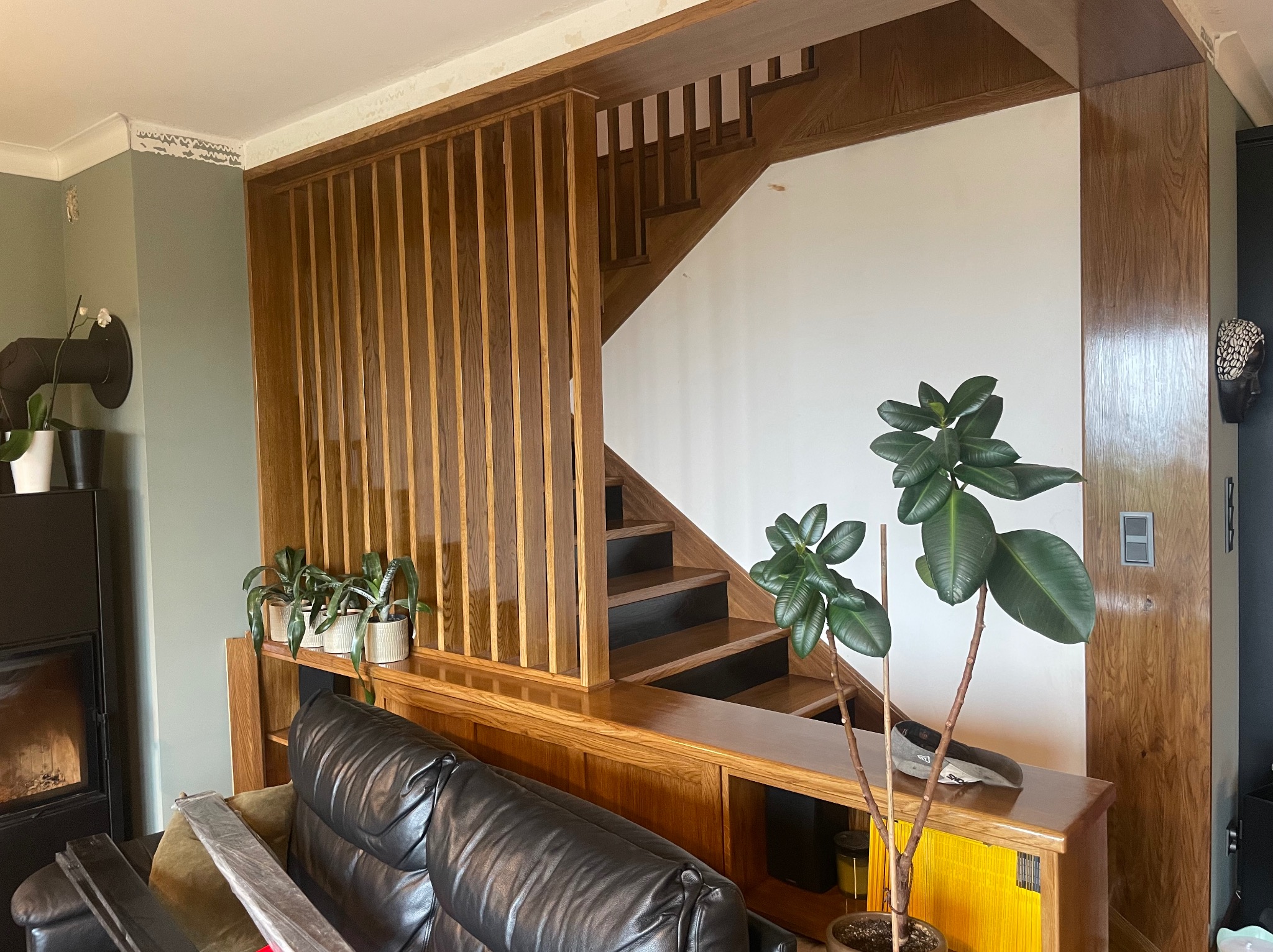 Drewniane schody z barierką i ażurową ścianką działową z pionowych listew. Elementy wykończenia wnętrza z drewna w ciepłym odcieniu brązu. Rośliny doniczkowe.