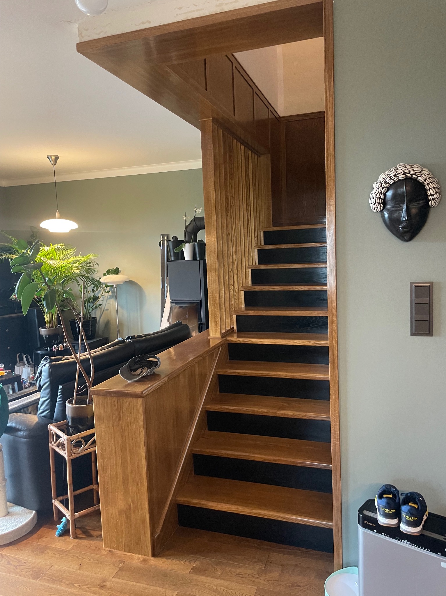 Drewniane schody z czarnymi podstopnicami, widoczne z dołu, z elementami wystroju wnętrza: rośliny, maska na ścianie, buty. Naturalne oświetlenie.