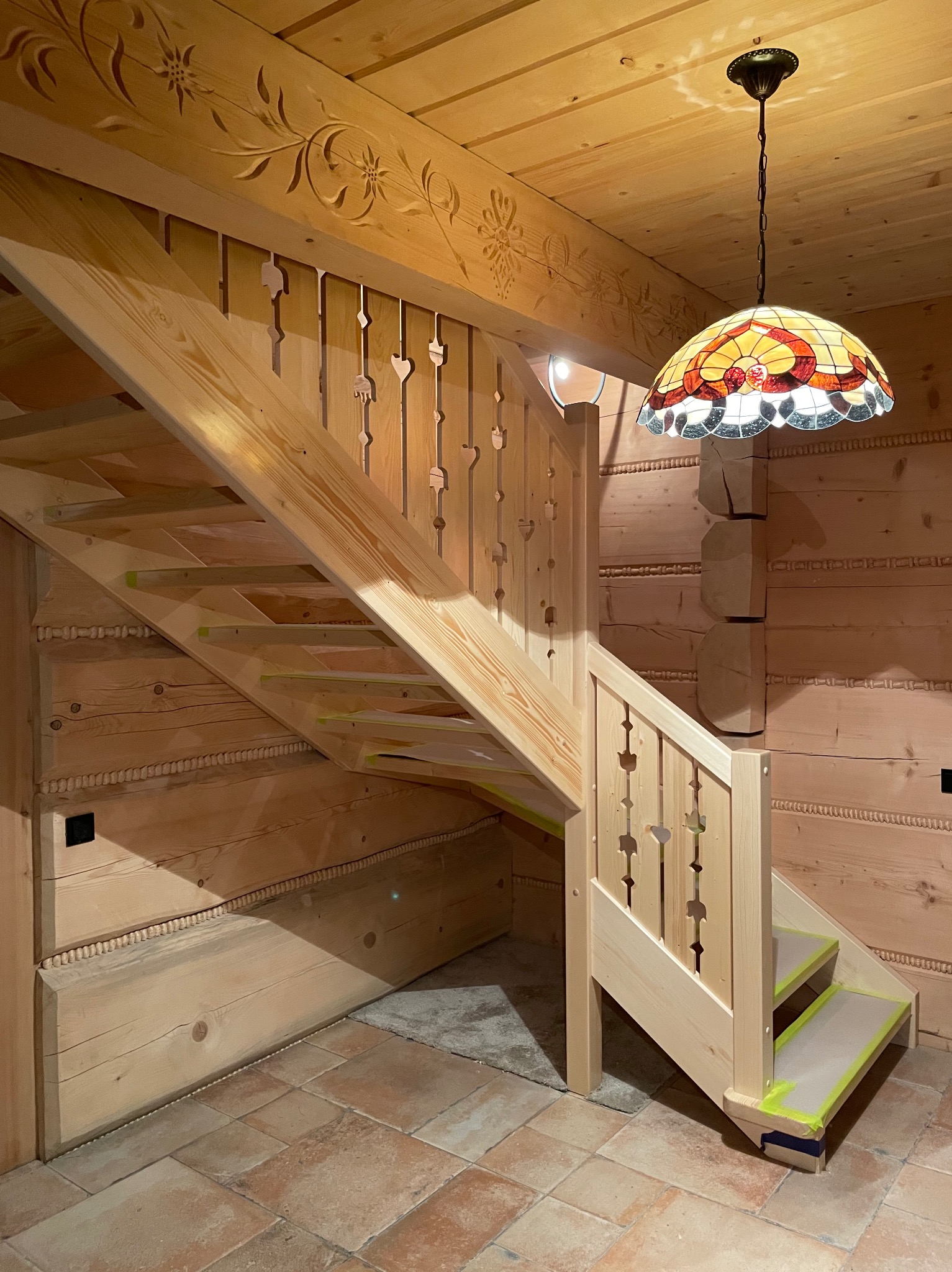 Drewniane schody z ozdobnymi balustradami w góralskim stylu, podświetlone lampą witrażową. Widoczne zdobienia na belkach stropowych.