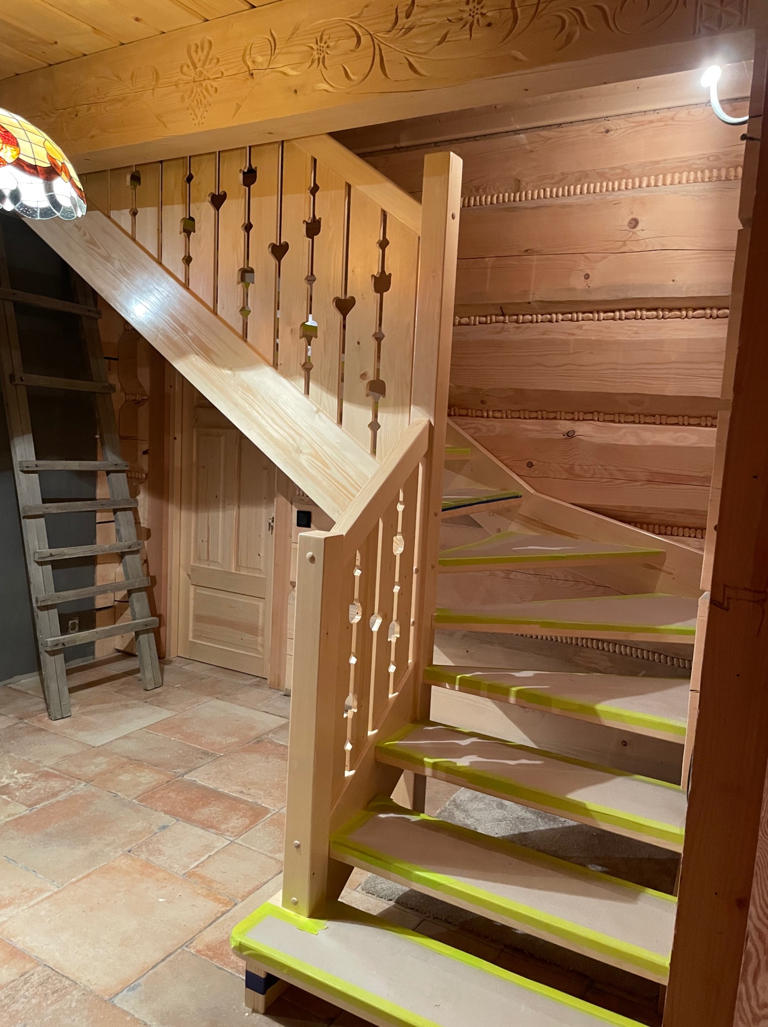Drewniane schody w stylu góralskim z balustradą z wyciętymi sercami, zabezpieczone taśmą malarską przed wykończeniem. W tle ściany i sufit z rzeźbionego drewna.