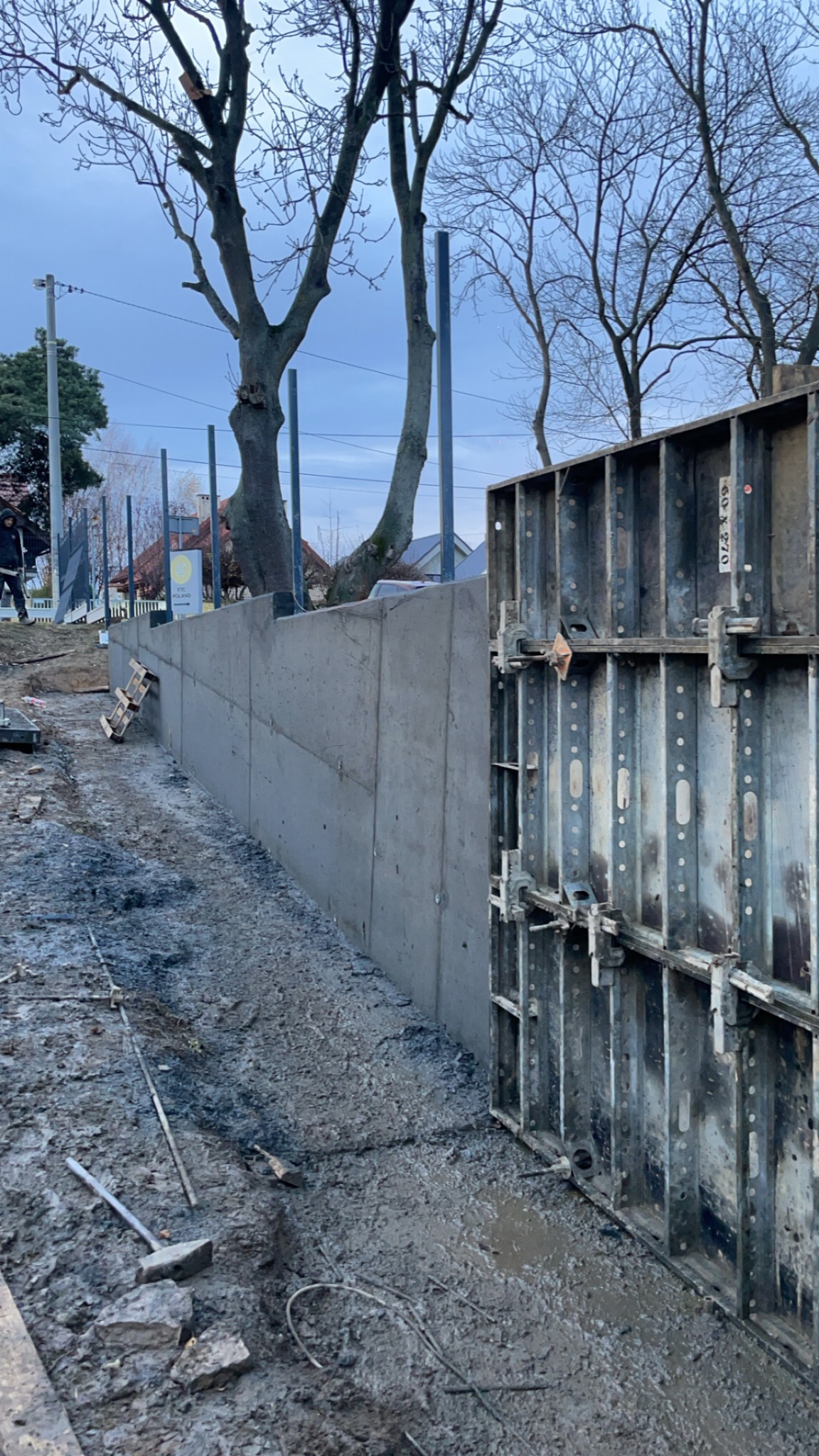 Betonowy mur oporowy w trakcie budowy, widoczne szalunki systemowe. W tle drzewa bez liści i fragment zabudowy. Na ziemi błoto i narzędzia budowlane.