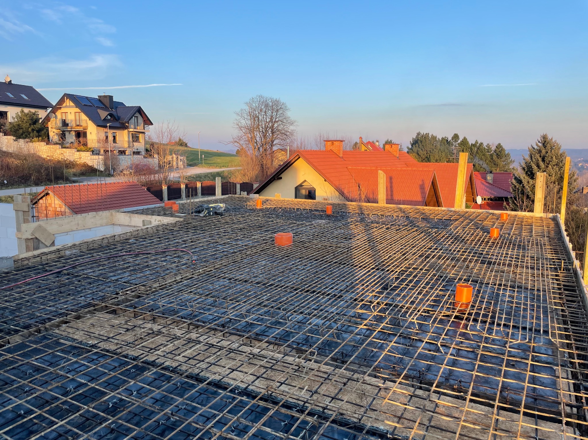 Zbrojenie stropu domu jednorodzinnego z widocznymi elementami instalacji i deskowaniem, w tle zabudowa mieszkalna i błękitne niebo o poranku.