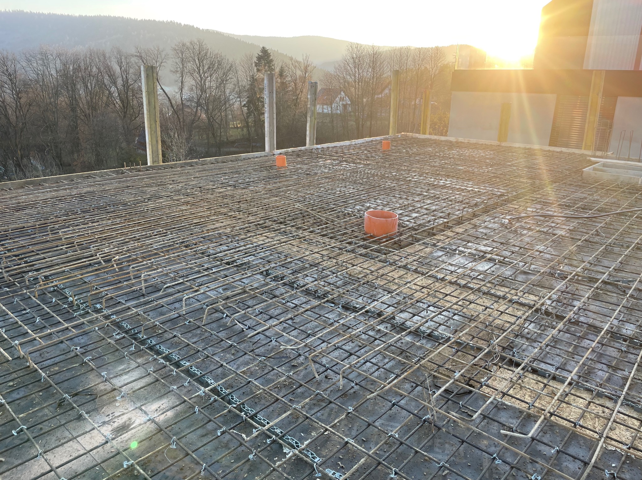 Zbrojenie płyty fundamentowej domu w trakcie budowy, widok z góry na siatkę z prętów stalowych, z pomarańczowymi rurami wentylacyjnymi. W tle zalesione wzgórza.