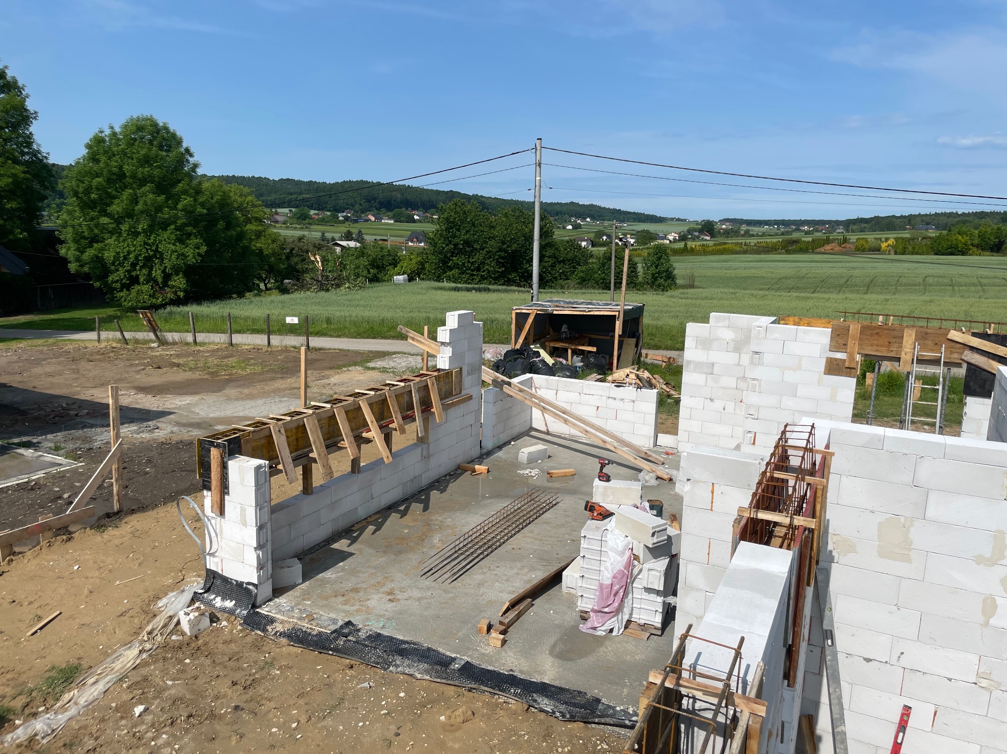 Budowa domu z białych bloczków: wznoszone ściany, szalunki drewniane, zbrojenie. W tle zielony krajobraz i błękitne niebo. Widok z góry na plac budowy.