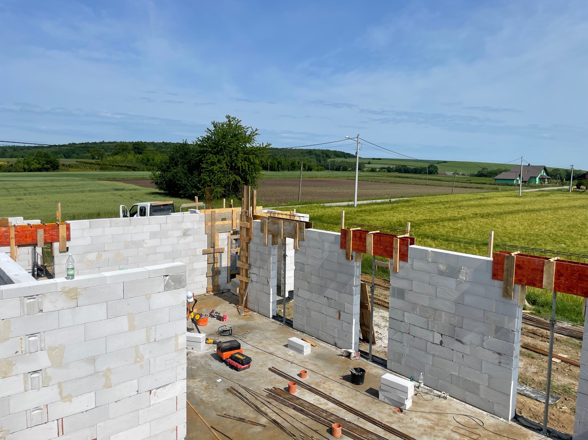 Wznoszenie ścian domu z betonu komórkowego. Widoczne narzędzia, zbrojenia i szalunki. W tle zielone pola i błękitne niebo. Ujęcie z góry.