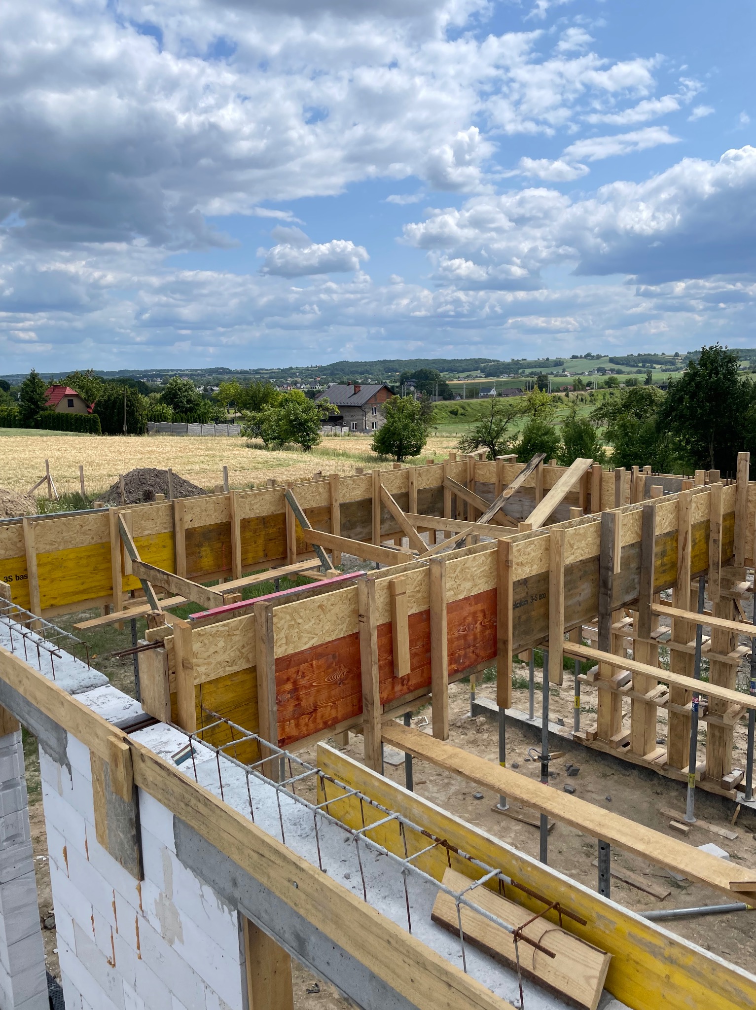 Budowa domu: szalunek fundamentów z drewnianych desek i stalowych podpór na tle krajobrazu. Widoczne zbrojenia i fragment ściany z białych bloczków.
