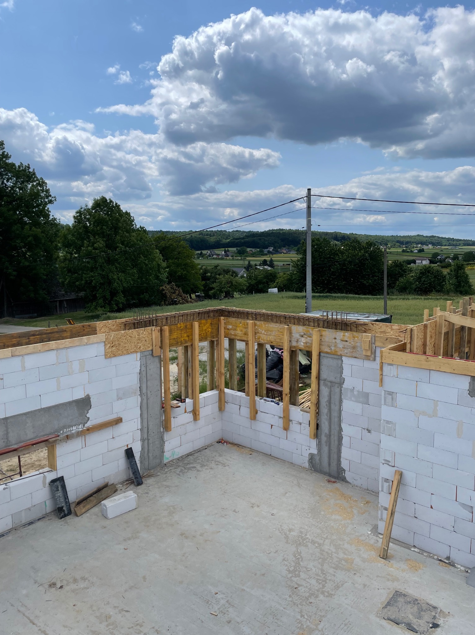Wnętrze domu w trakcie budowy: białe ściany z betonu komórkowego, drewniane szalunki okienne i stropowe, widok na krajobraz z błękitnym niebem i chmurami.