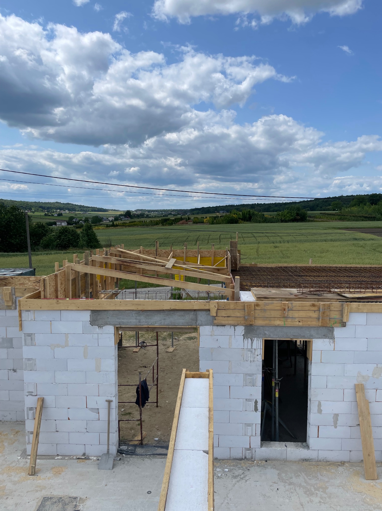 Wznoszenie ścian domu z betonu komórkowego, widoczne szalunki i zbrojenia stropu. W tle zielone pola i błękitne niebo z chmurami.