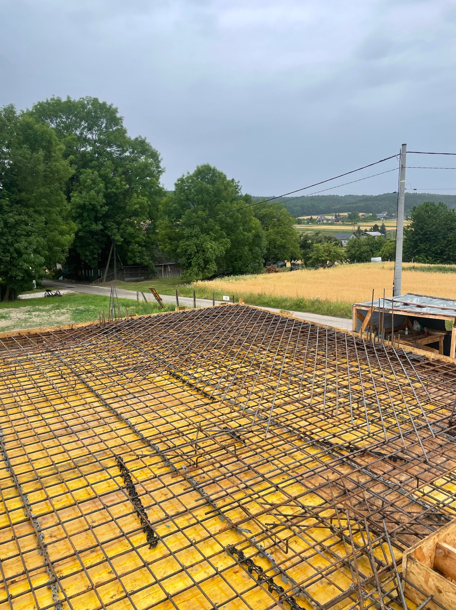 Zbrojenie fundamentu na żółtym szalunku, widok z góry na tle zieleni i pola. Elementy konstrukcyjne przygotowane do zalania betonem, etap budowy domu.