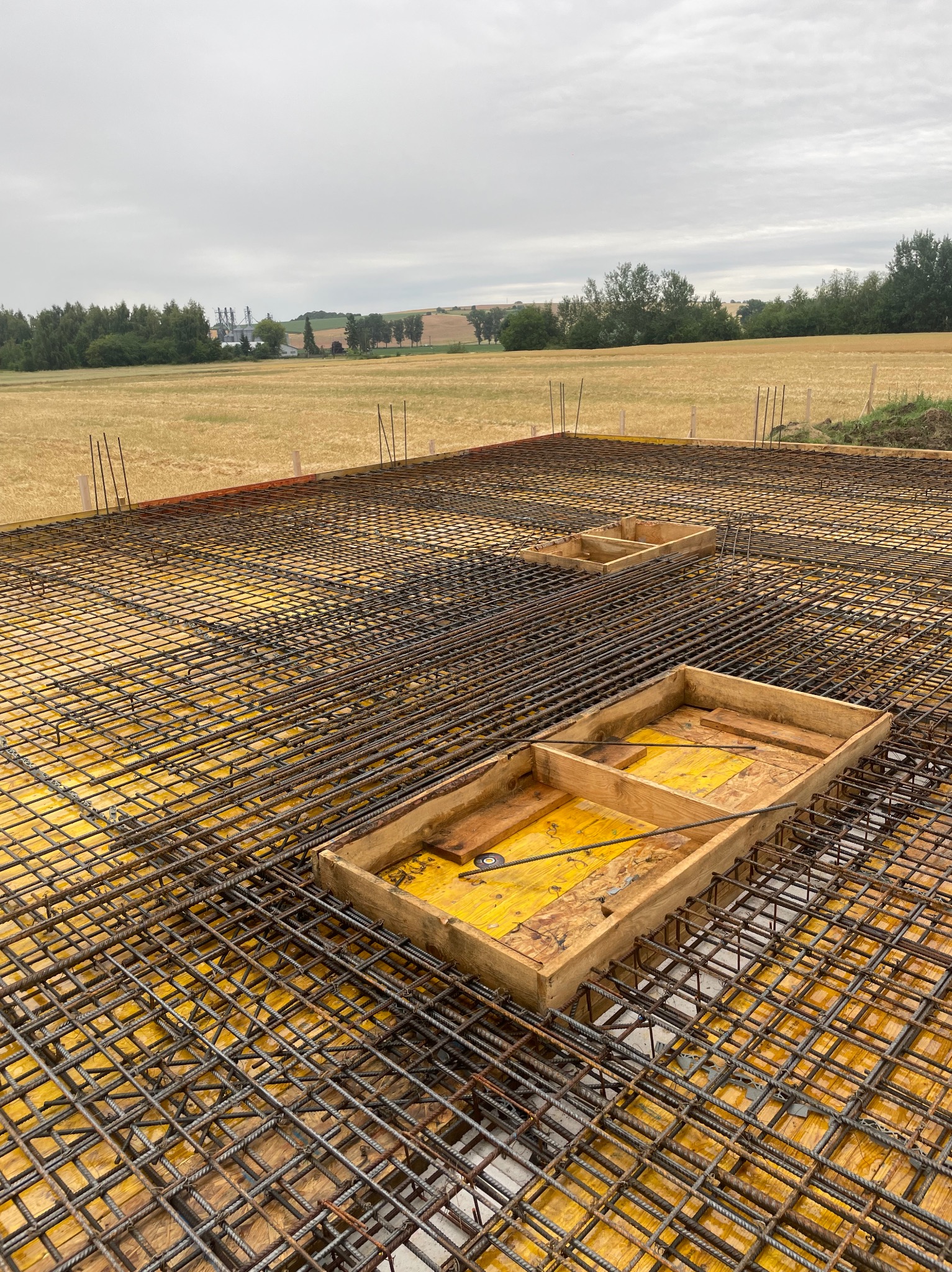 Zbrojenie fundamentu z żółtą szalunką i drewnianymi formami na tle pola i pochmurnego nieba. Widok z góry ukazuje siatkę zbrojeniową i detale konstrukcyjne.
