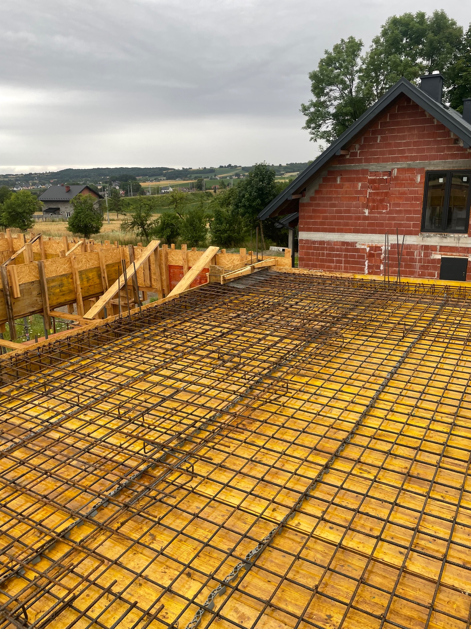 Zbrojenie stropu domu jednorodzinnego z czerwonej cegły, widok z góry na siatkę zbrojeniową na żółtym szalunku, w tle zielone wzgórza i pochmurne niebo.
