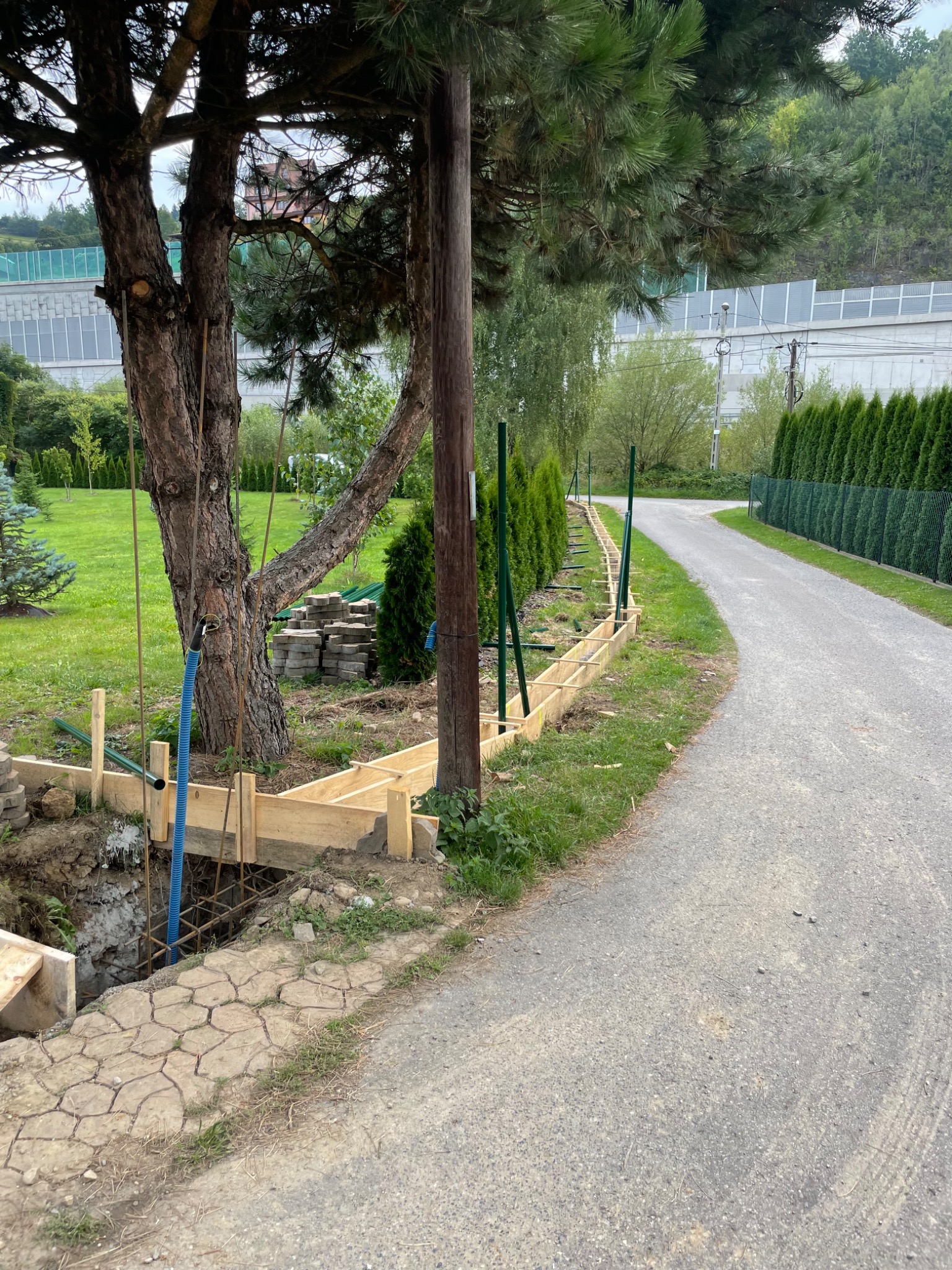Wykop pod fundament z drewnianym szalunkiem i zbrojeniem, widoczne ułożone obok kostki brukowe. W tle zielony trawnik, drzewa i droga.