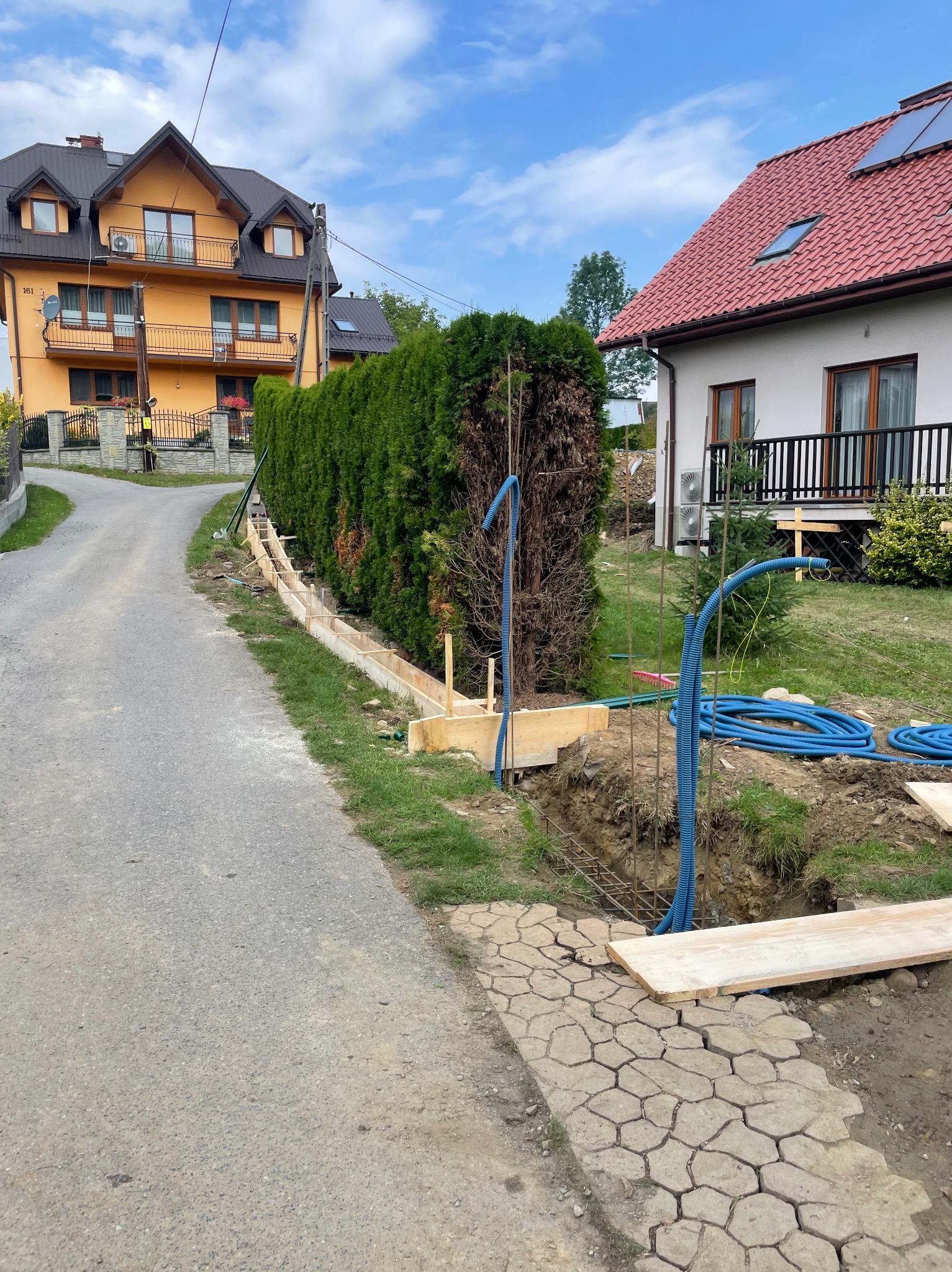 Przygotowanie do wylania fundamentów: szalunek z desek, zbrojenie i niebieskie rury instalacyjne w wykopie, na tle zielonego żywopłotu i dwóch domów.