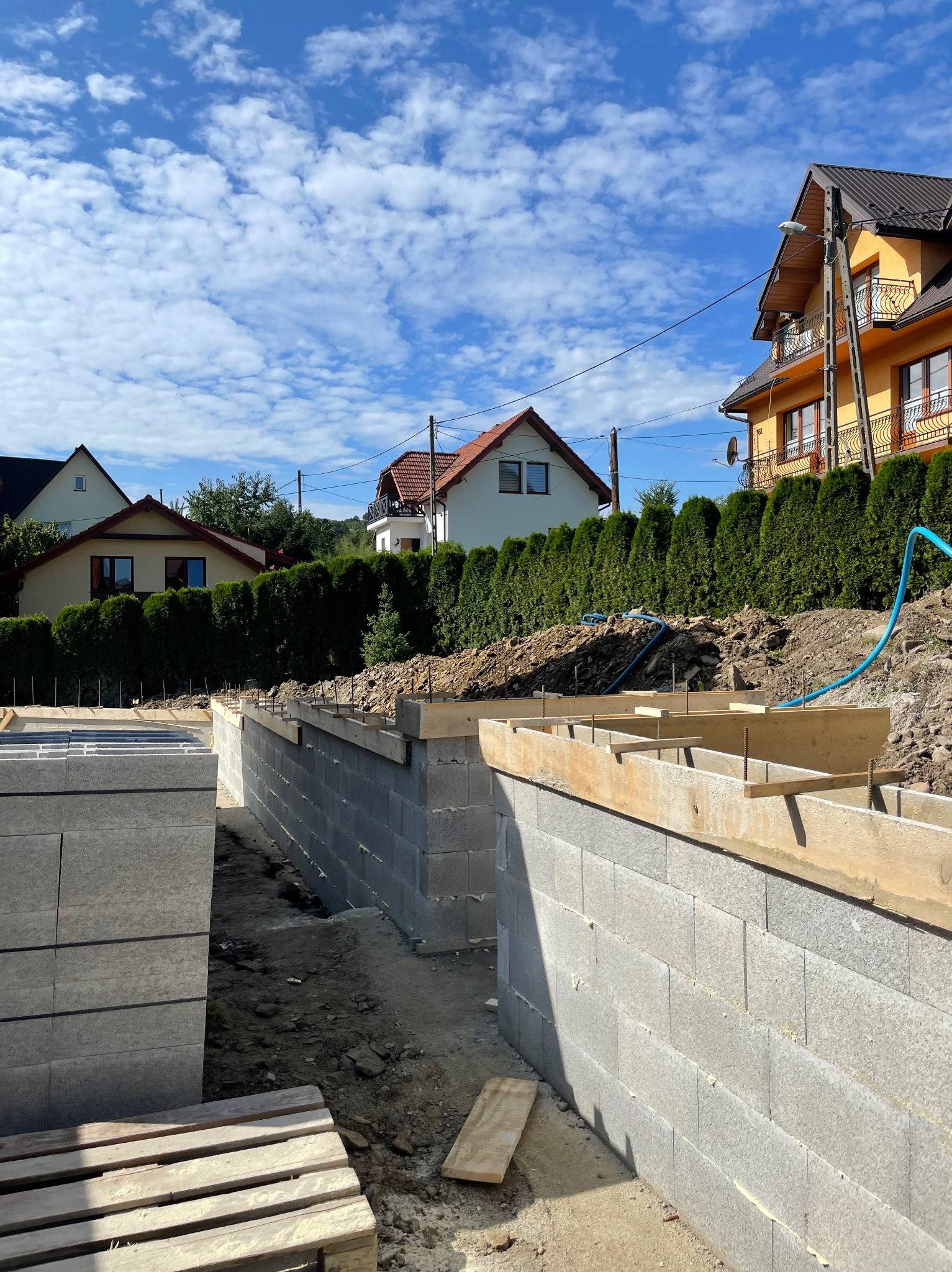 Wznoszenie fundamentów z bloczków betonowych na tle zabudowy jednorodzinnej i błękitnego nieba z chmurami. Widoczne szalunki i zbrojenie.