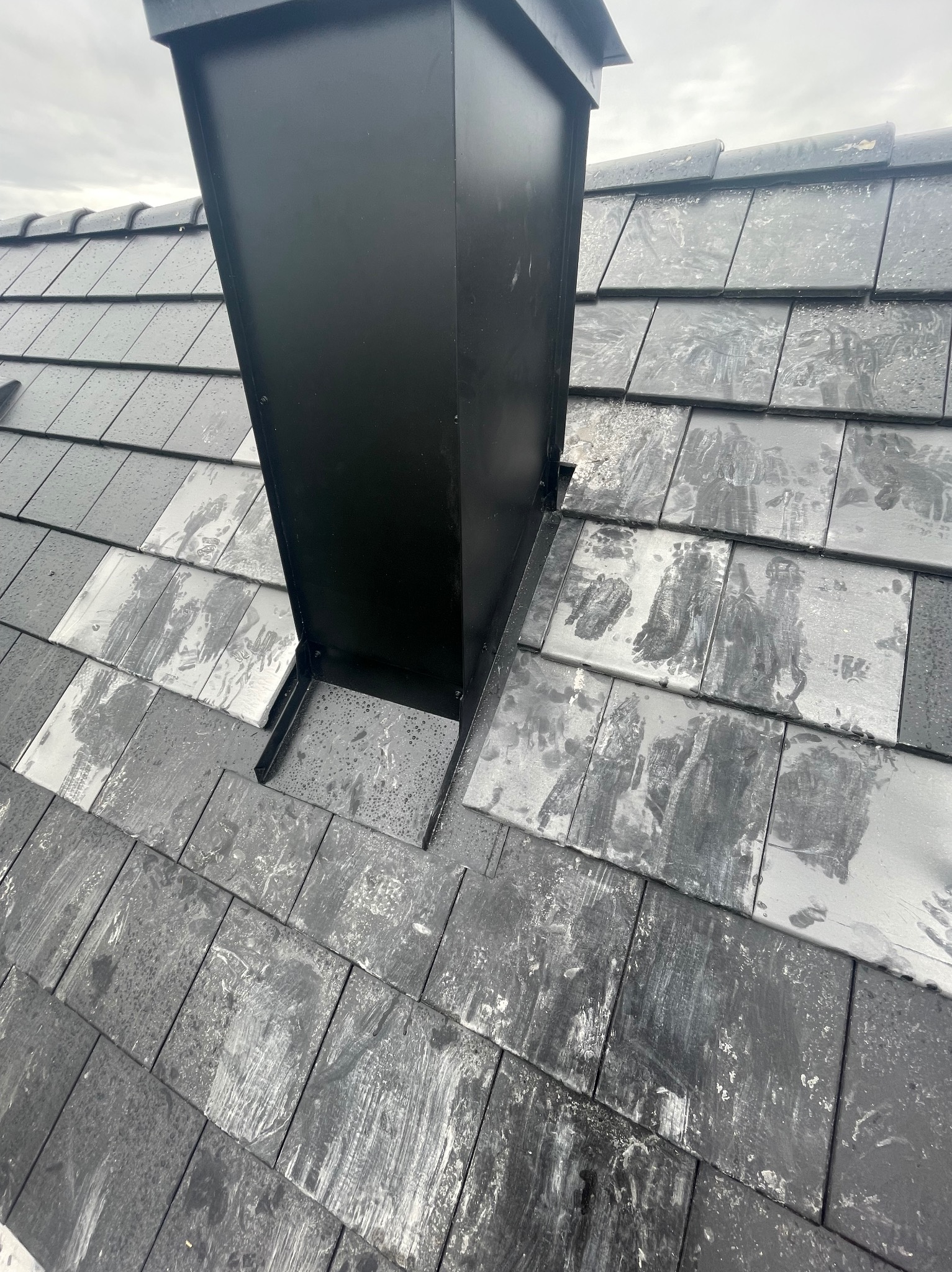 Szczegółowe ujęcie dachu z czarną, metalową obudową komina, wykonanego z ciemnych, kwadratowych dachówek pokrytych plamami. Widok z góry, pochmurne niebo w tle.