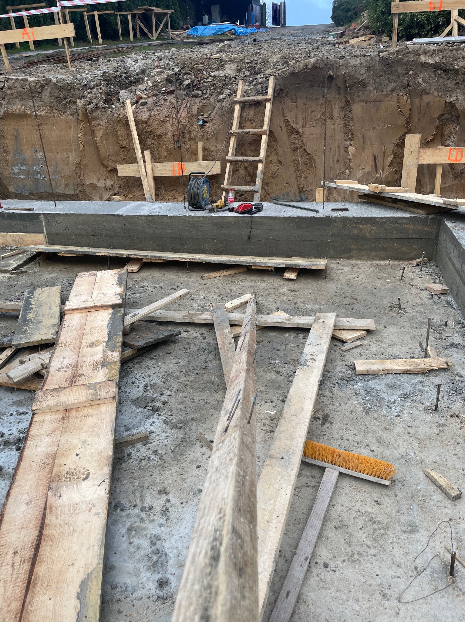 Wykop pod fundamenty z częściowo wylanym betonem, widoczne szalunki, drabina oparta o ziemię, narzędzia i kable. Etap budowy w trakcie realizacji.